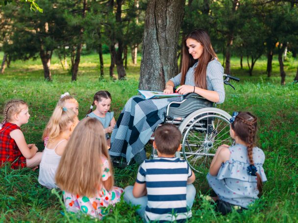 Eine junge Frau im Rollstuhl liest einer Gruppe von Kindern aus einem Buch vor. Die Kinder sitzen auf einer Wiese um die junge Frau herum und hören aufmerksam zu.